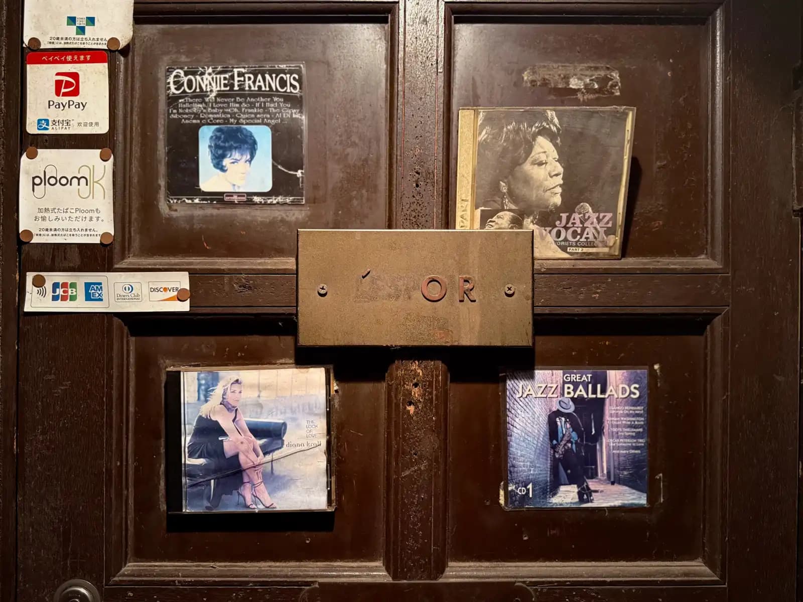 A dark wooden bar door decorated with old jazz CD covers, including Connie Francis and Diana Krall, next to payment stickers.