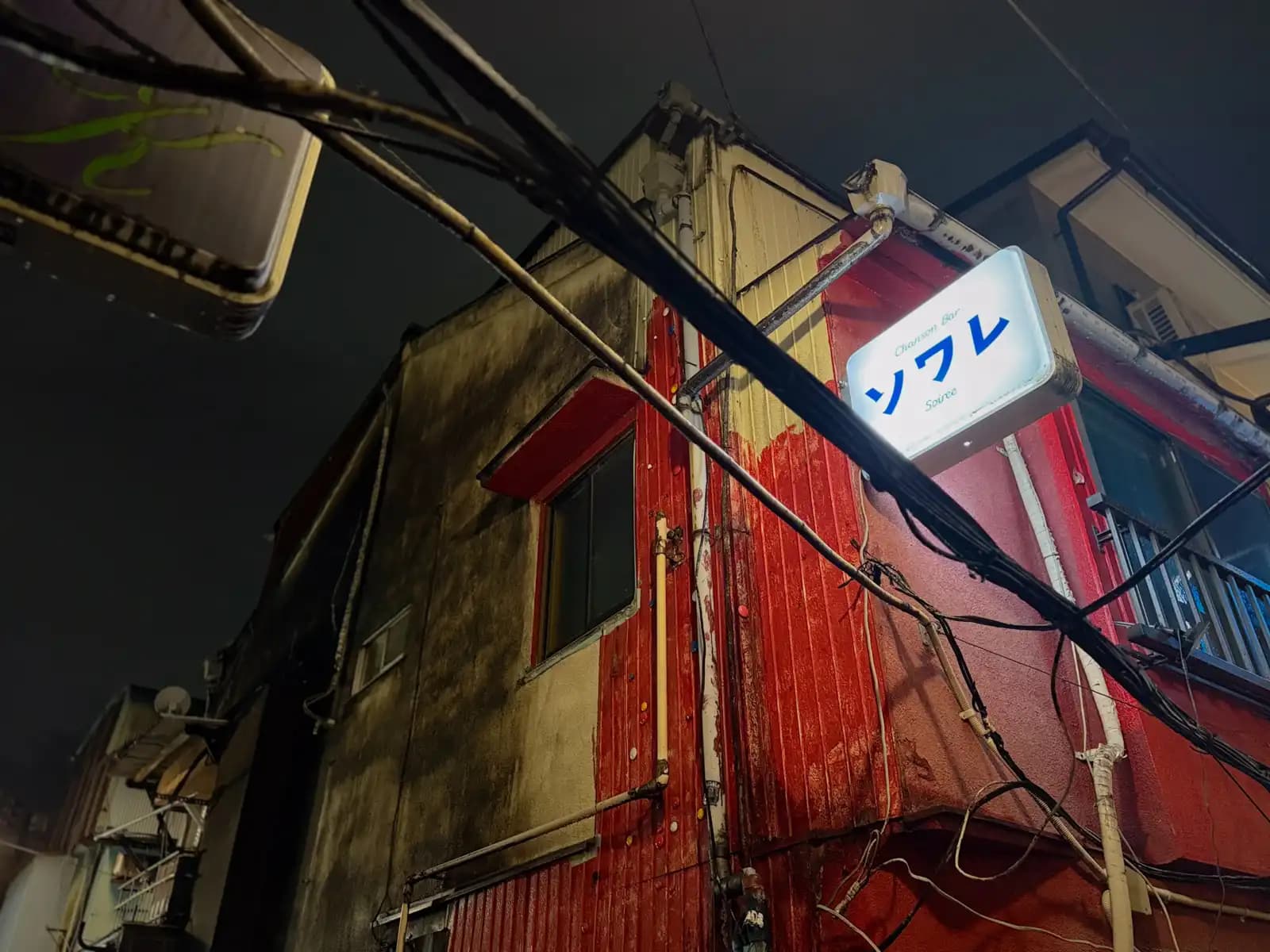 The lit sign for Chanson Bar Soirée on the corner of a worn building with a red-painted wall and wires running past.