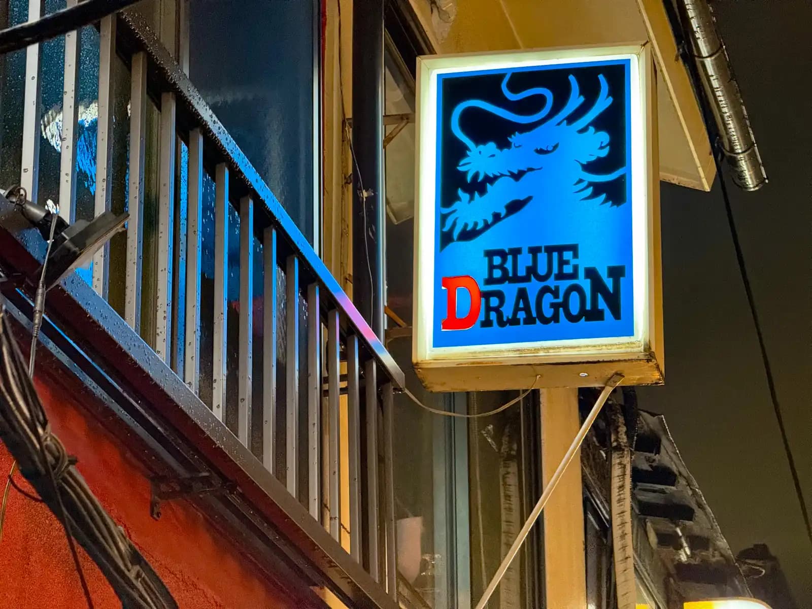 The lit Blue Dragon sign next to a second-floor railing still wet with raindrops.