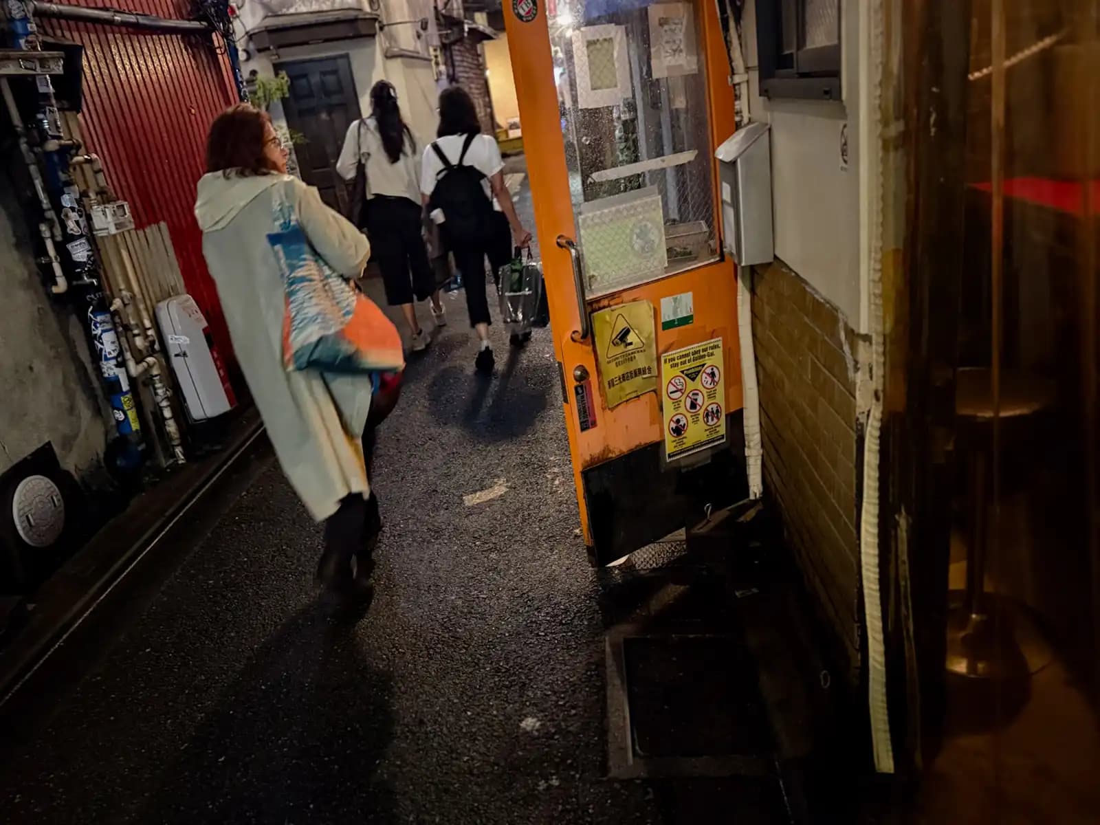 People walking single file down a tight alley, past an orange door with a sign saying if you can't follow the rules, stay out of Golden Gai.