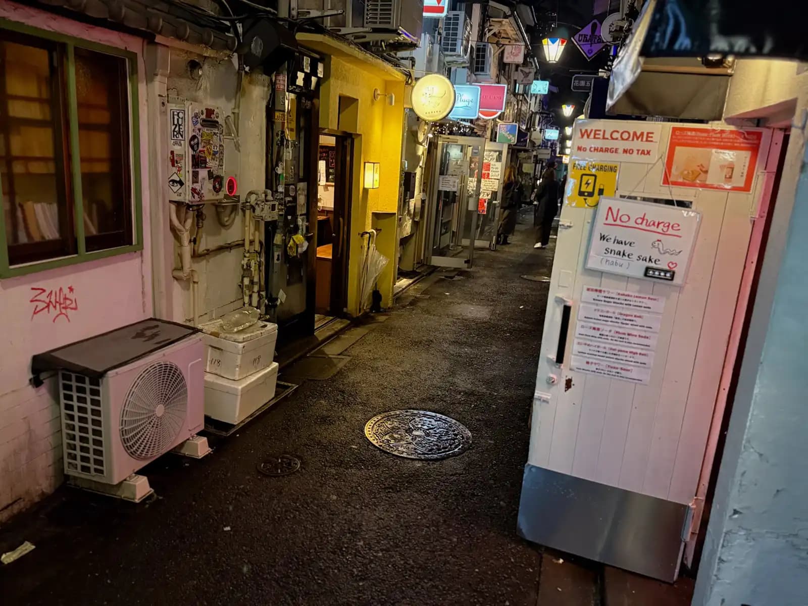 A white bar door with signs for no cover charge, free phone charging and habu snake sake, on a wet alley with a manhole cover.