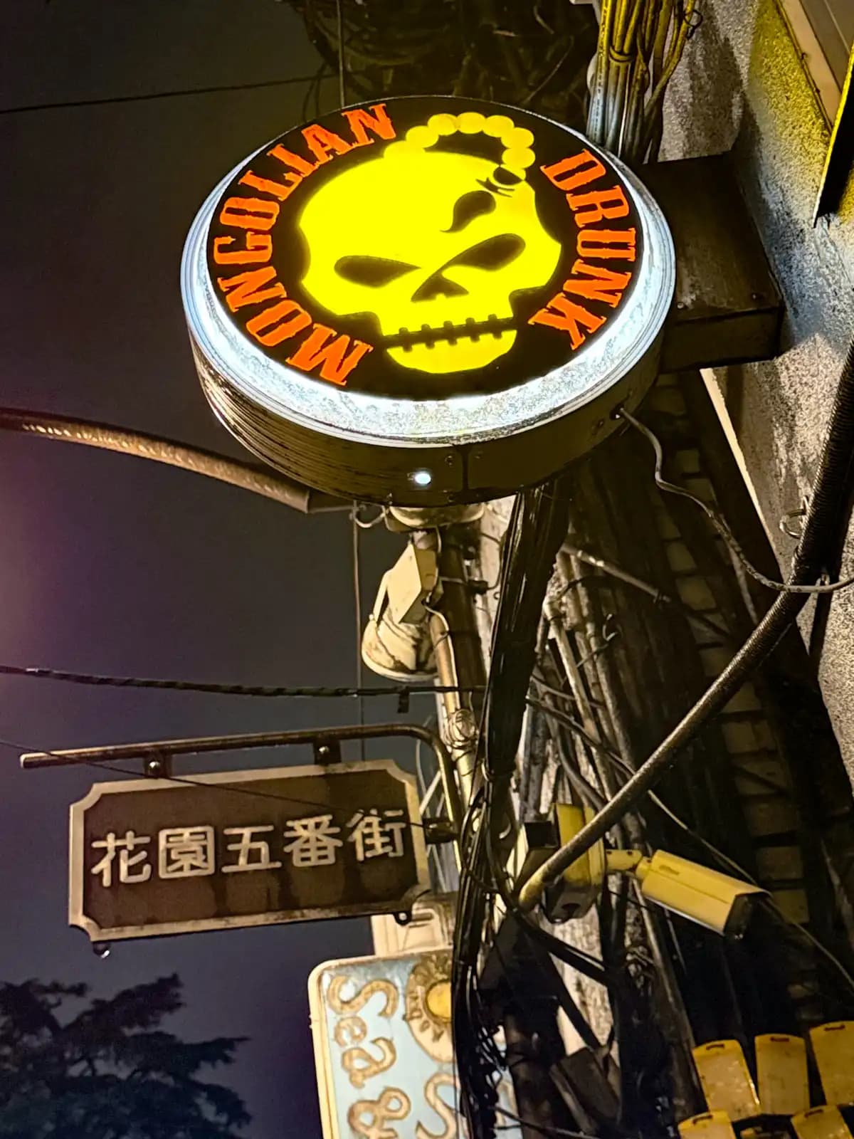 The round Mongolian Drunk sign with a yellow skull, hanging above the Hanazono Gobangai street sign.