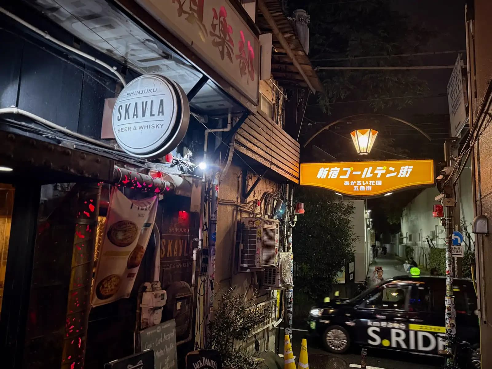 The Skavla beer and whisky bar on the corner, with the yellow Shinjuku Golden Gai arch and a taxi driving past on the street.