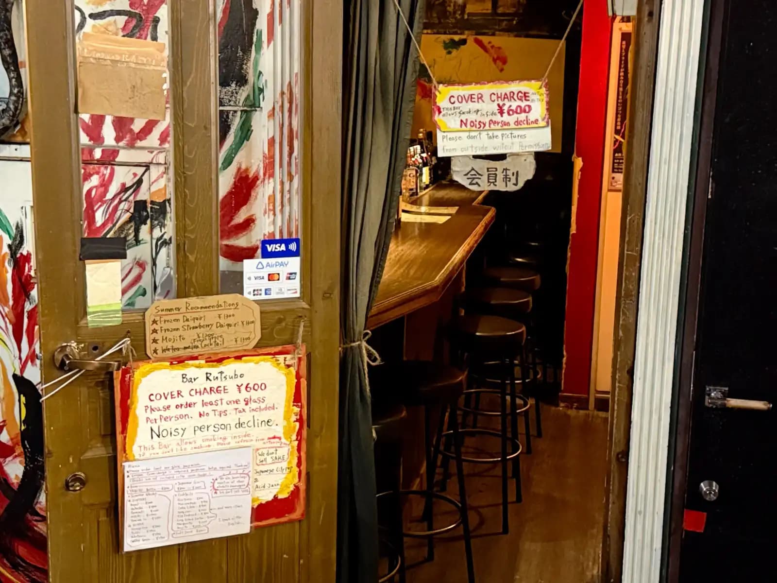 The open door of Bar Rutsubo, with handwritten signs for a 600 yen cover charge and "Noisy person decline," and a row of empty stools inside.