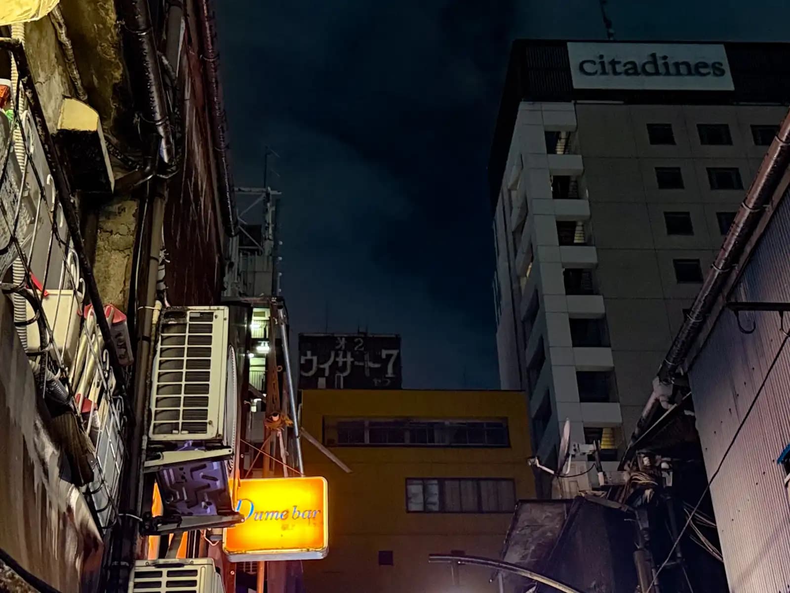 Looking up between buildings at the Citadines hotel, with an orange Dame bar sign lit up below.