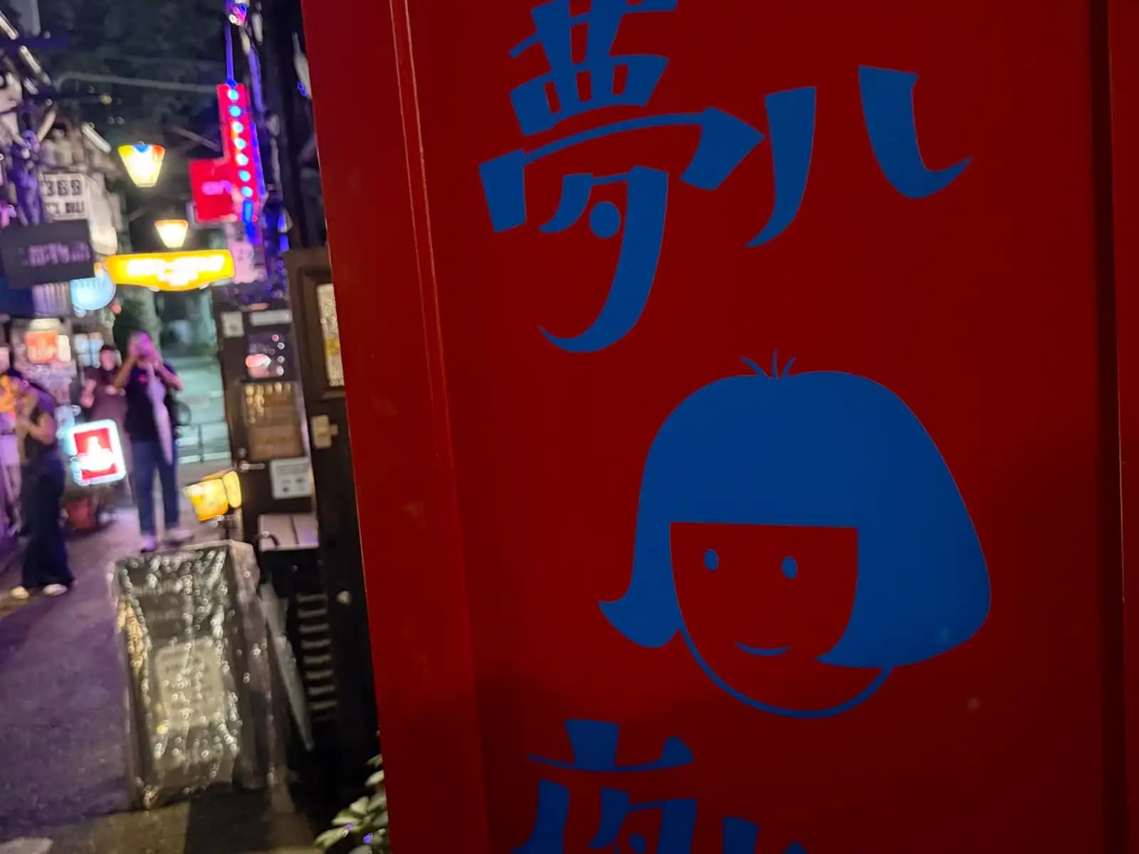 A red bar sign with a cartoon girl with a bob haircut, while people behind it stop to take photos in the alley.