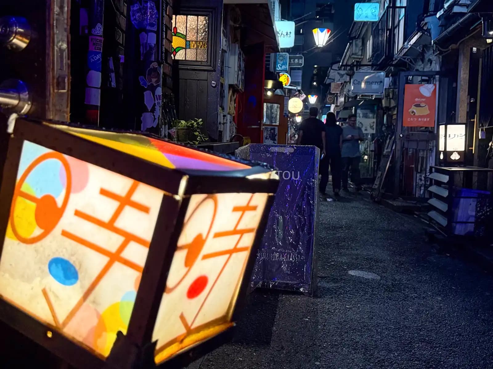 A glowing box lantern with a colorful pattern sits on the ground at the edge of a dark lane, with people chatting further down.
