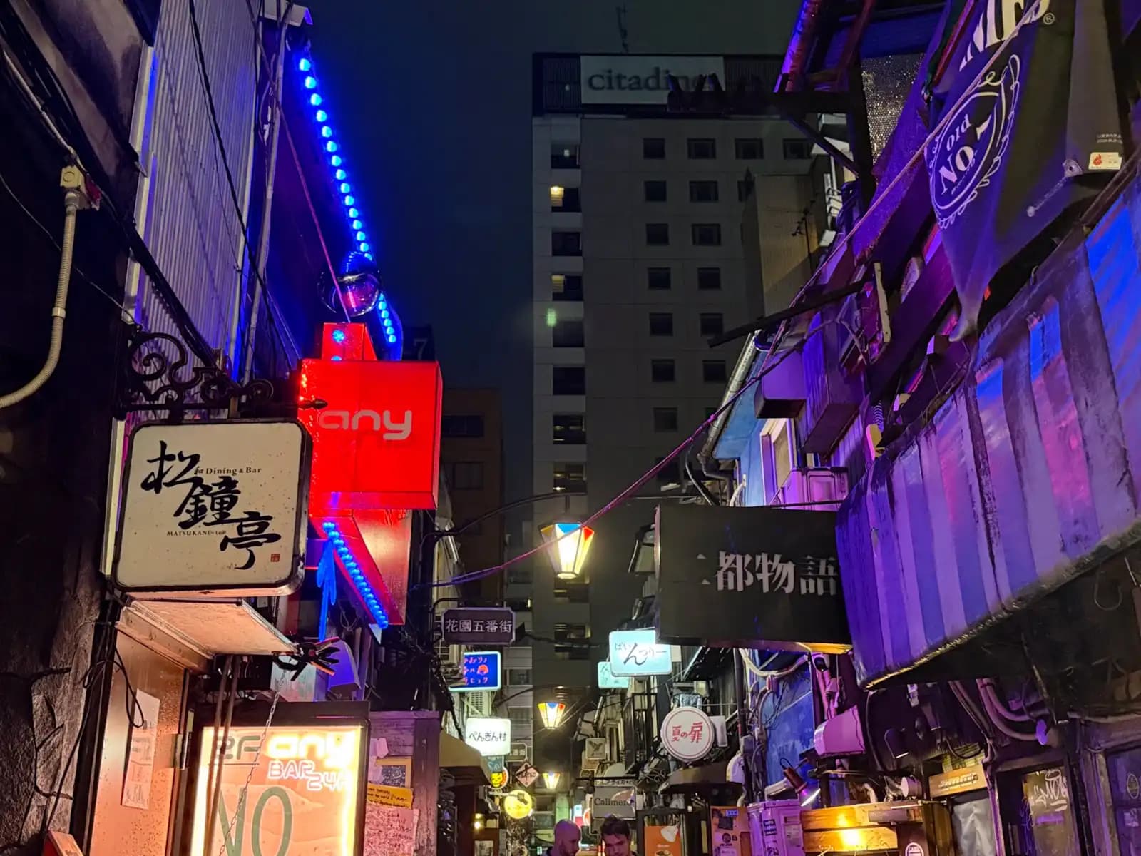 Signs for Matsukane-tei, a red one reading "any" and Nito Monogatari fill the Hanazono Gobangai lane, with the Citadines hotel rising behind.