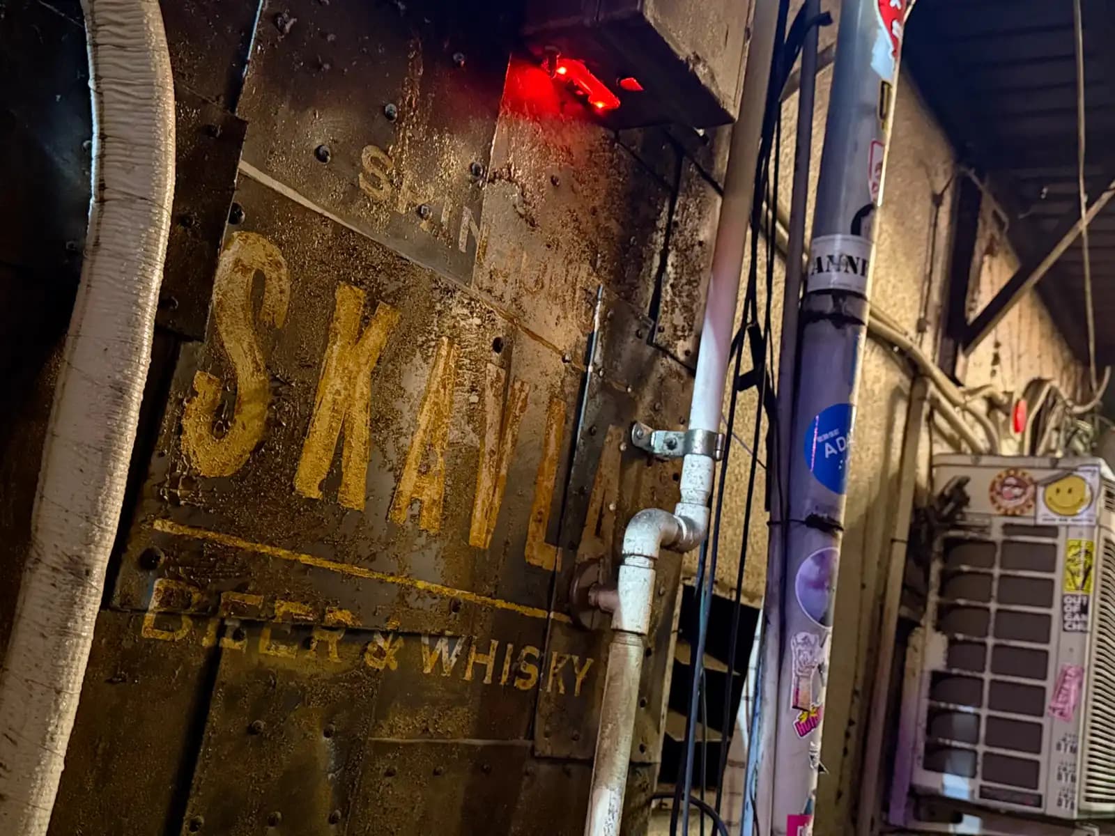 Faded yellow lettering for Shinjuku Skavla Beer & Whisky painted on a grimy metal wall, next to a pipe and a stickered air conditioner.