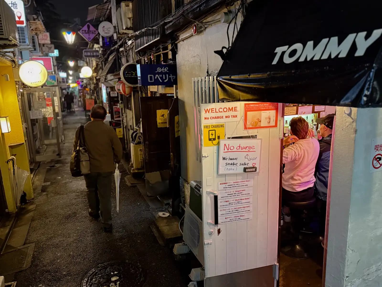 A man with an umbrella walks past the open door of a tiny bar under a Tommy awning, where a couple of customers sit at the counter.