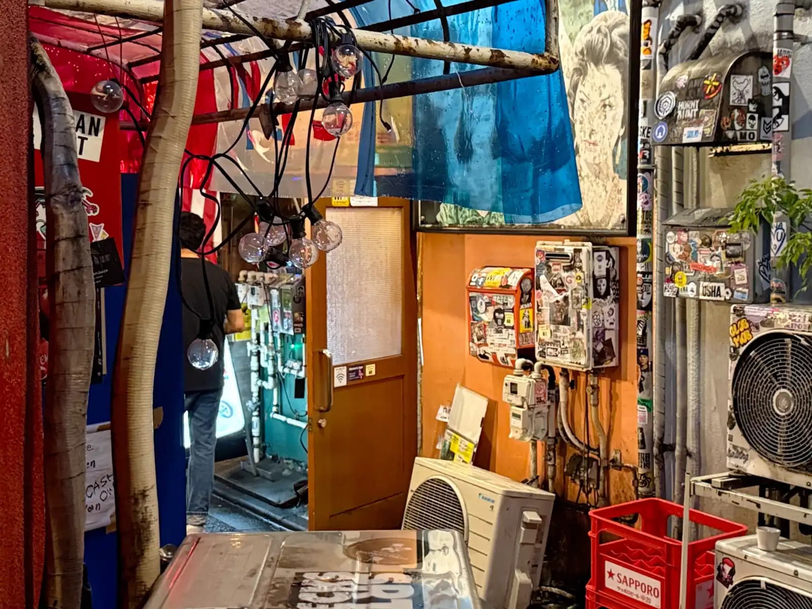 A cramped back corner full of sticker-covered meter boxes, AC units, a Sapporo beer crate and string lights.