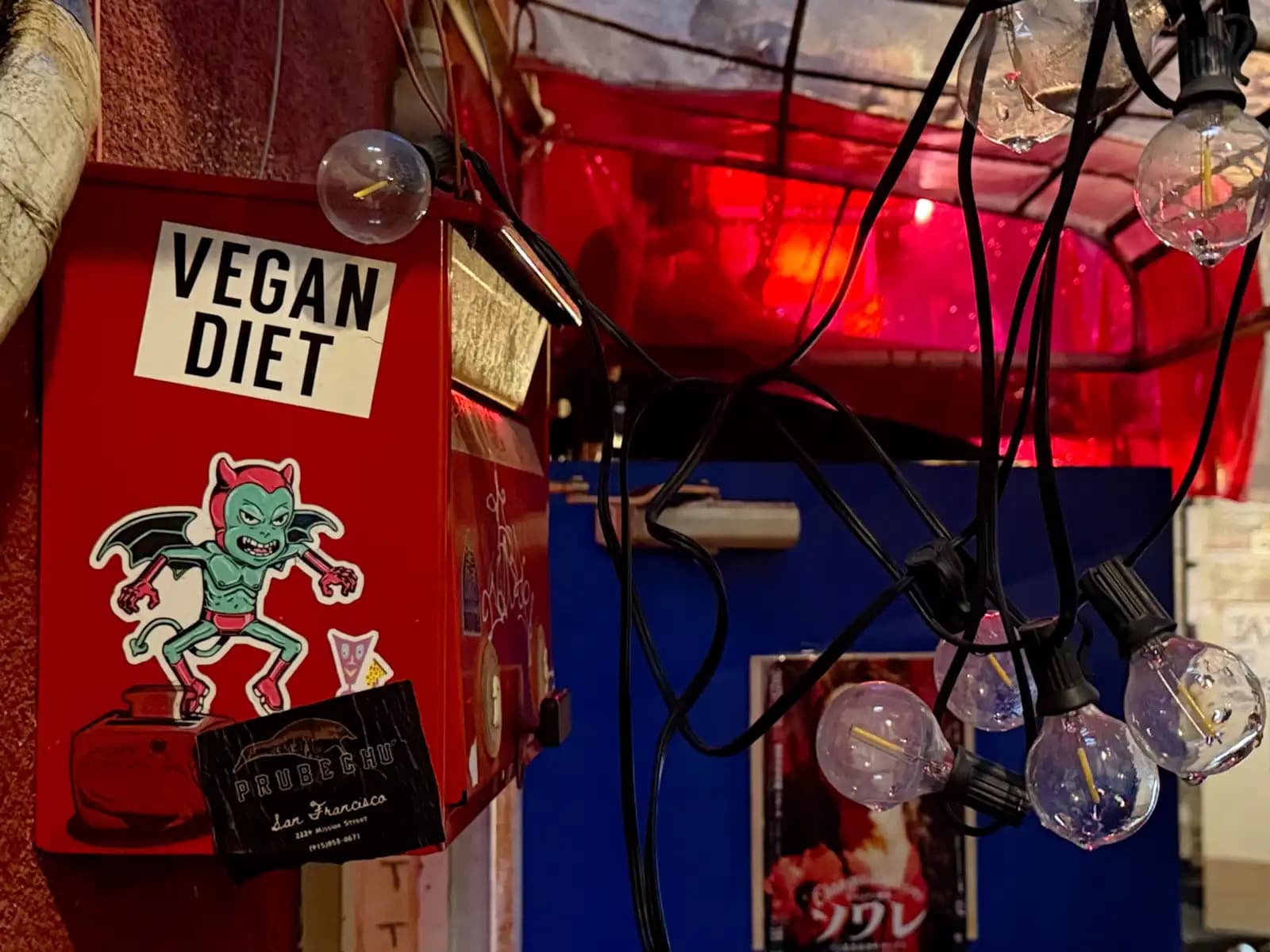 A red box by a bar door covered in stickers, including one that says "Vegan Diet" and a green devil.