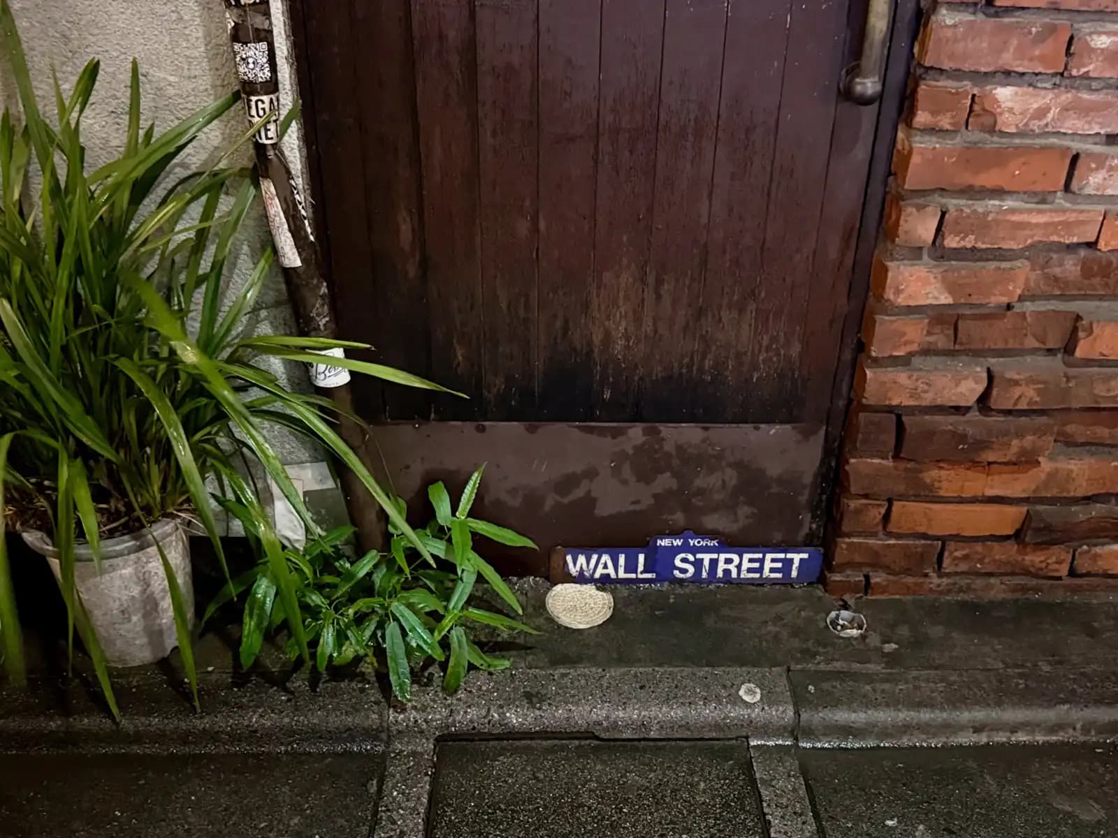 A small "Wall Street" sign leaning against a worn wooden door, next to potted plants and a brick wall.