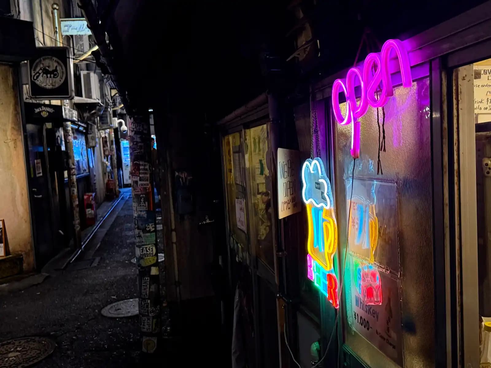 A pink neon "open" sign and other neon signs in a bar window, with a dark, wet alley running off to the left.