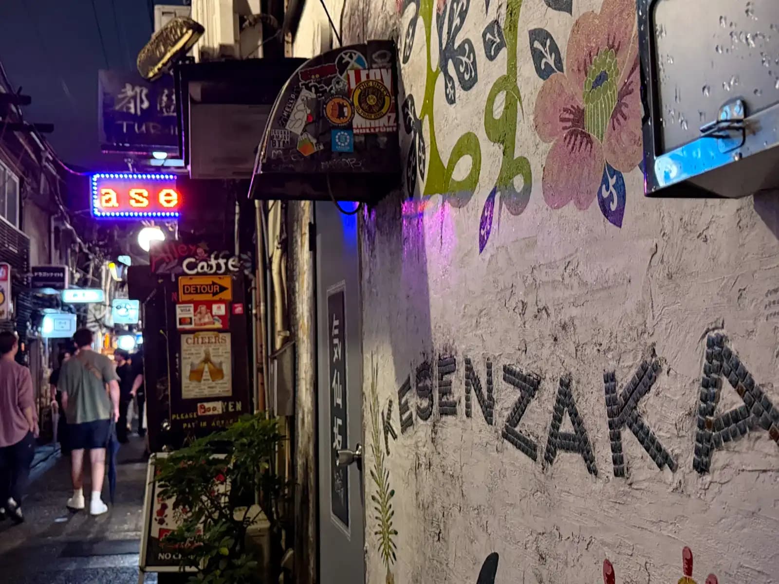 A bar name spelled out in black tiles on a painted wall with big flowers, as people walk down the alley behind.