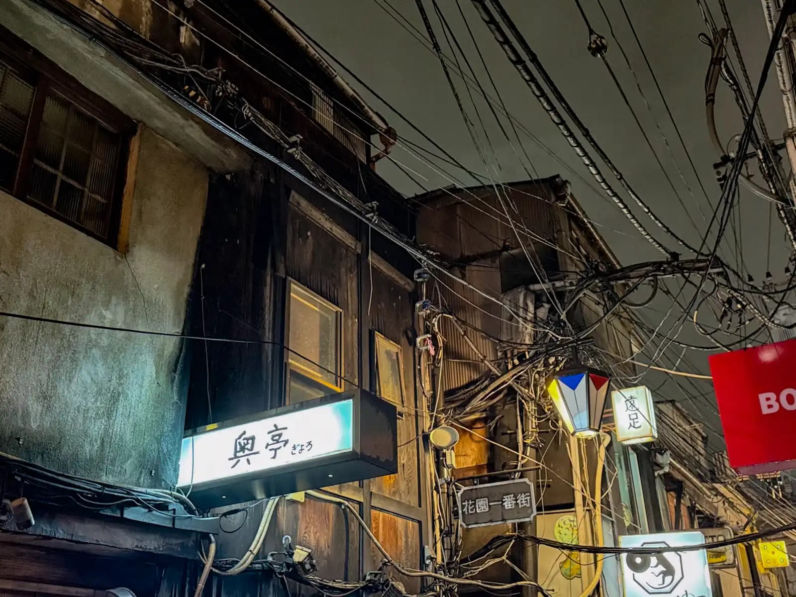 Old wooden buildings with a mess of power lines overhead and the lit sign for Gyoro above a doorway.