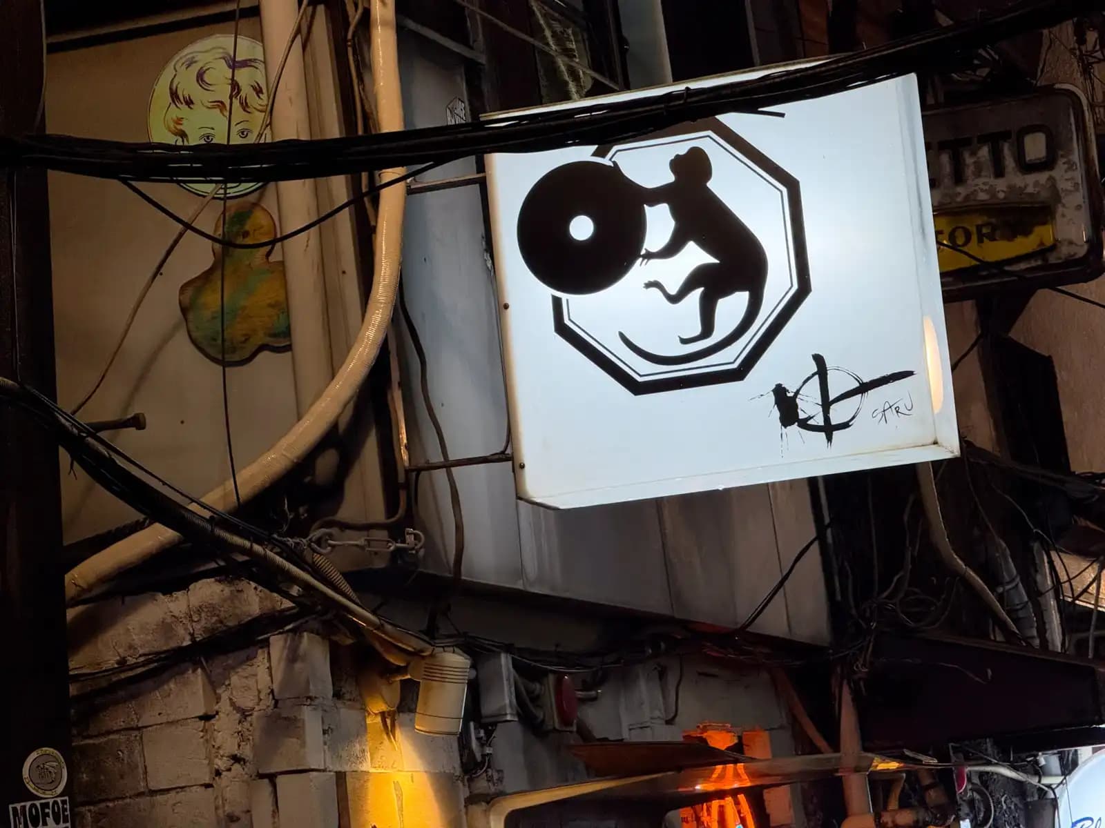 A lit sign with a monkey hugging a vinyl record, hanging among tangled cables and old pipes.