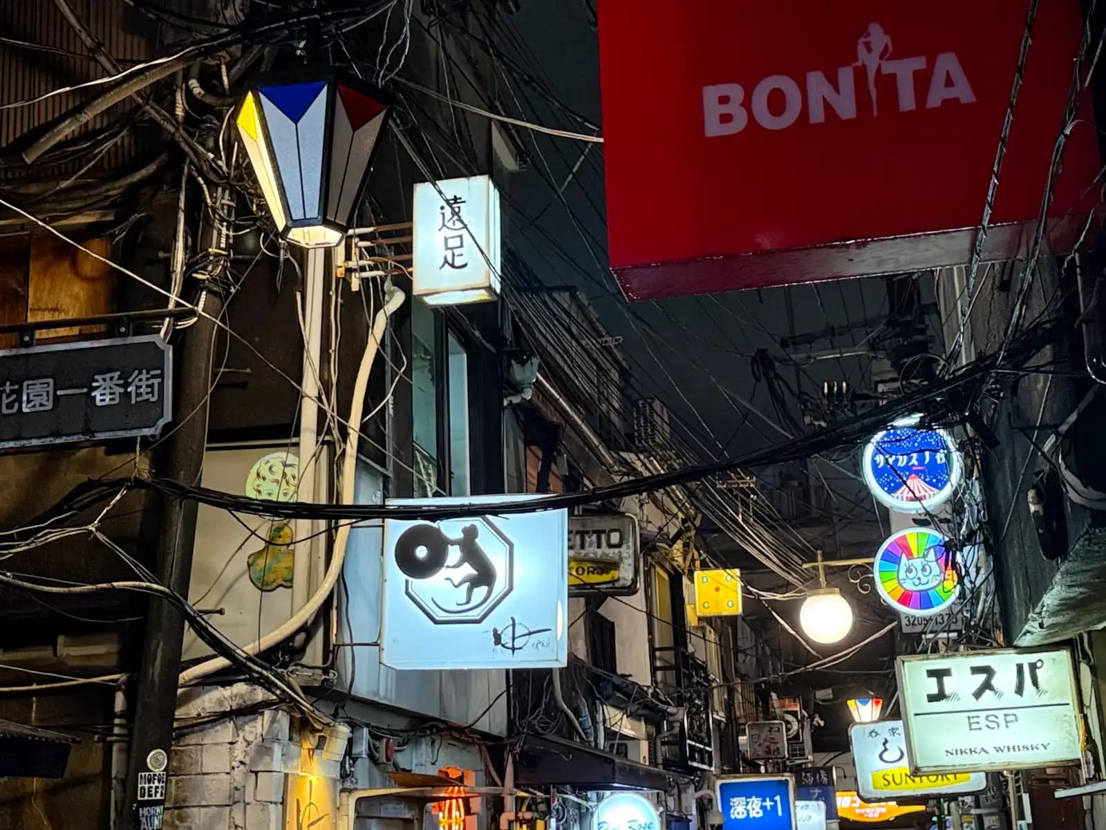 Signs stacked up along the alley: Bonita, Ensoku, ESP and a rainbow cat, under a web of wires.