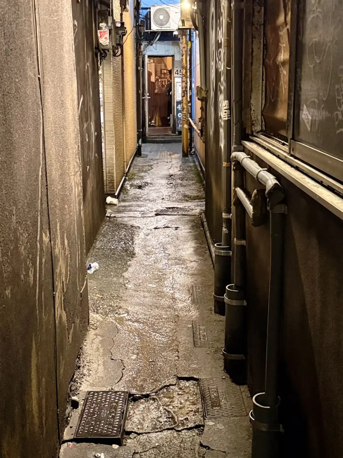 A passage barely wide enough for one person, with wet cracked concrete, drainpipes and a lit doorway at the far end.