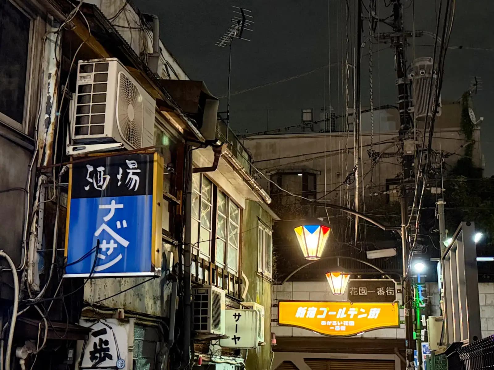 The blue Nabesan sign on an old building, with the Shinjuku Golden Gai sign glowing under lantern-shaped street lamps.
