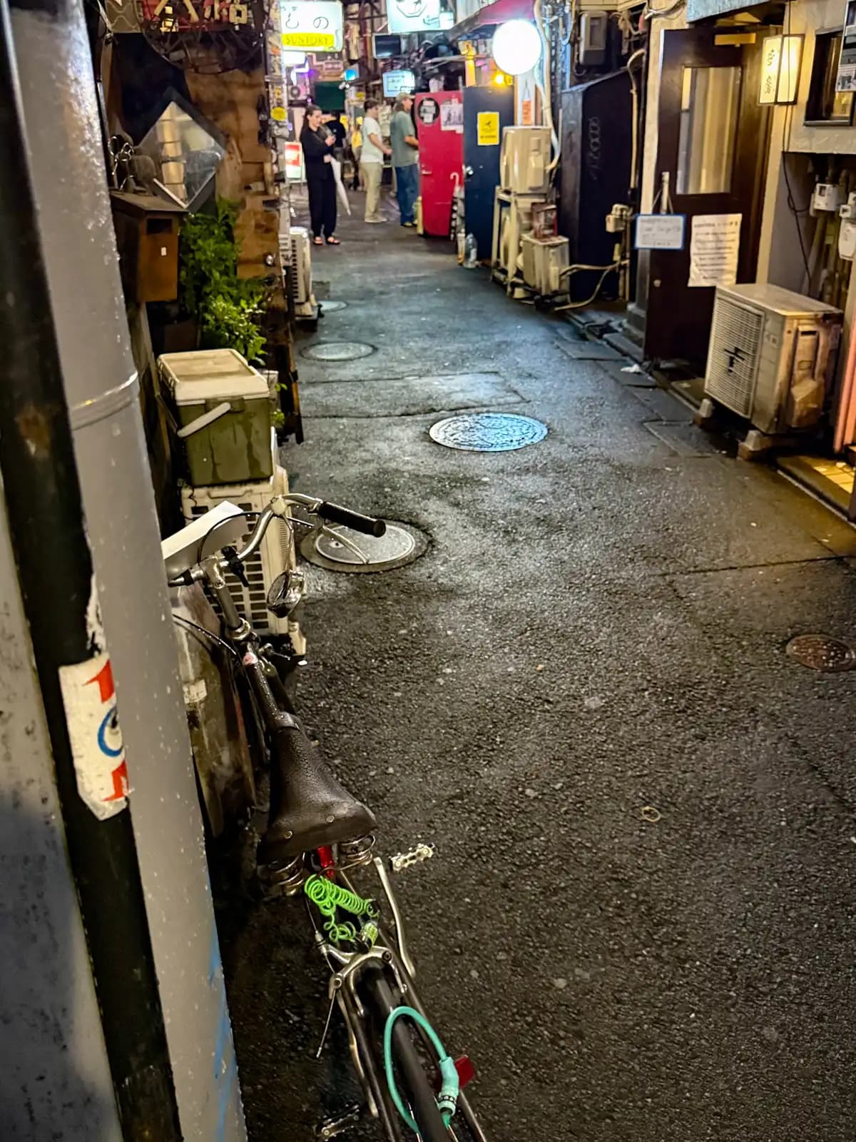 A bike parked against a pole in a wet alley, with a few people standing around outside bars farther down.