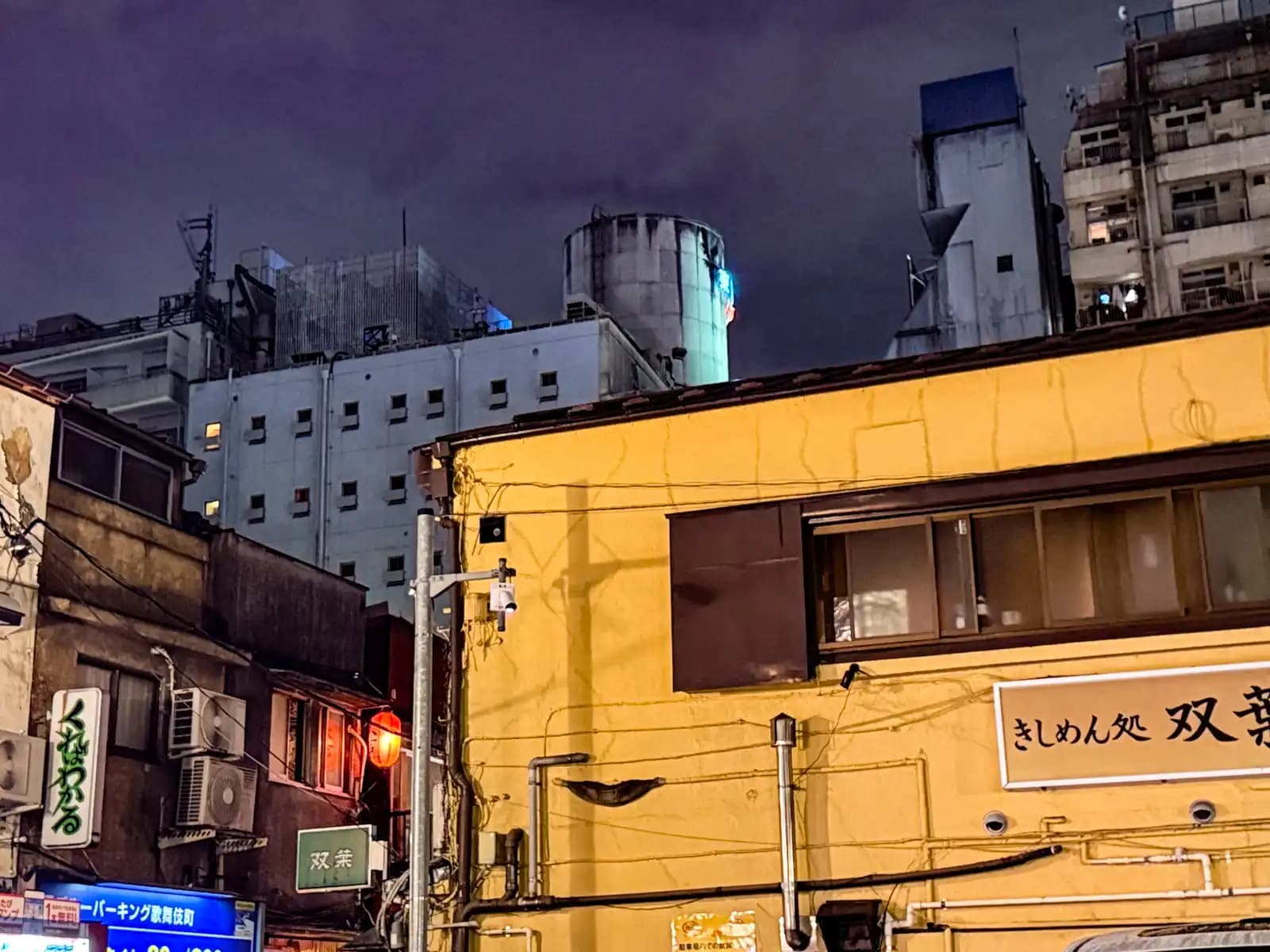 A yellow building with a kishimen noodle shop sign, and taller concrete buildings behind it under a cloudy night sky.