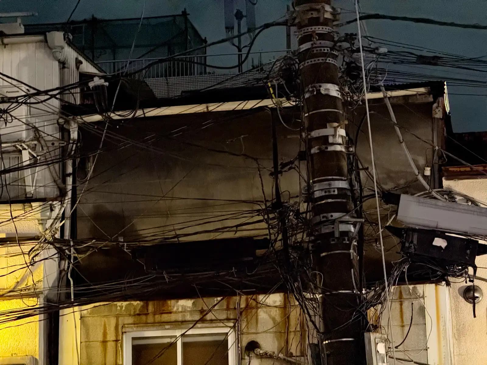 A utility pole with a huge tangle of wires and cables running in every direction in front of a dark building.