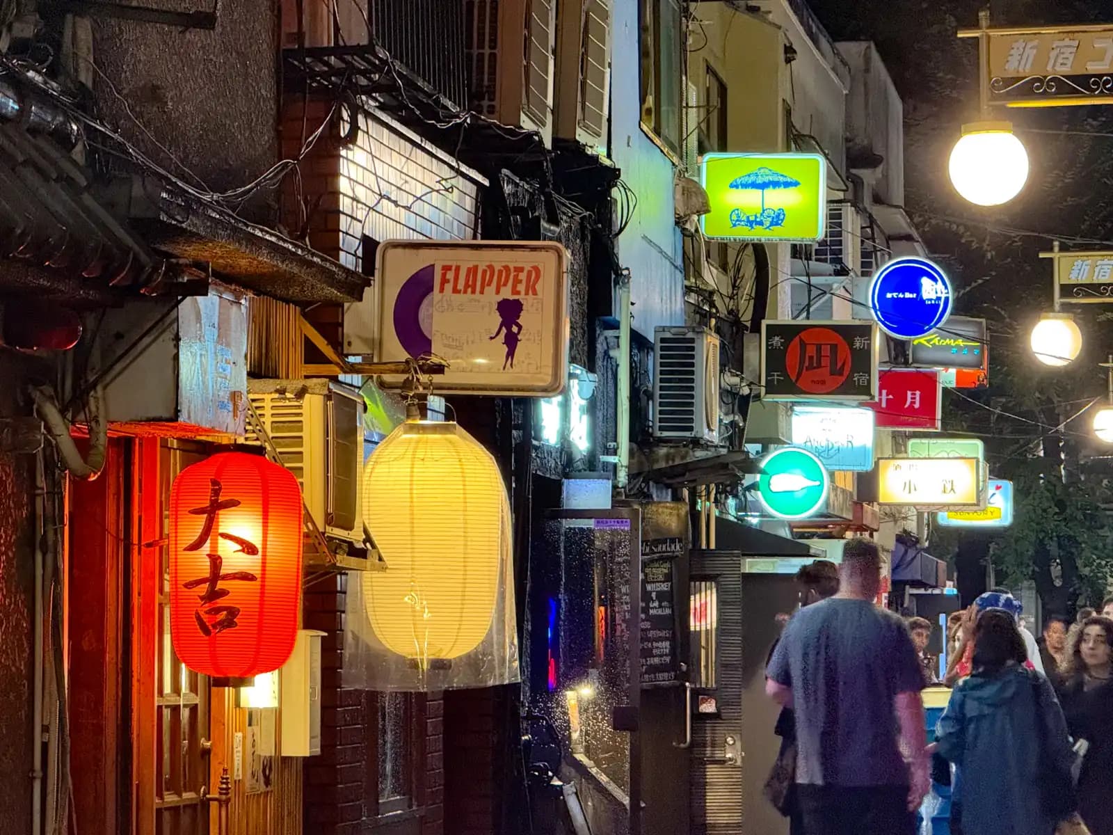 A red lantern for Daikichi and a white one under the Flapper sign, with people walking by and more bar signs down the street.