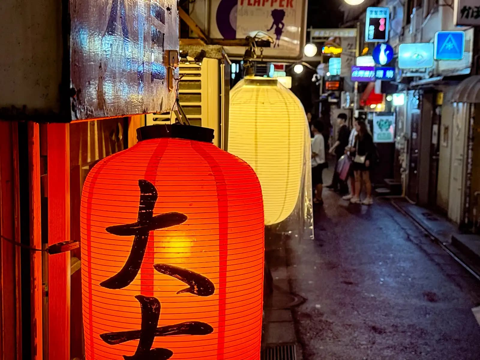 Up close to the red Daikichi lantern, with a white lantern next to it and people chatting in the wet street.
