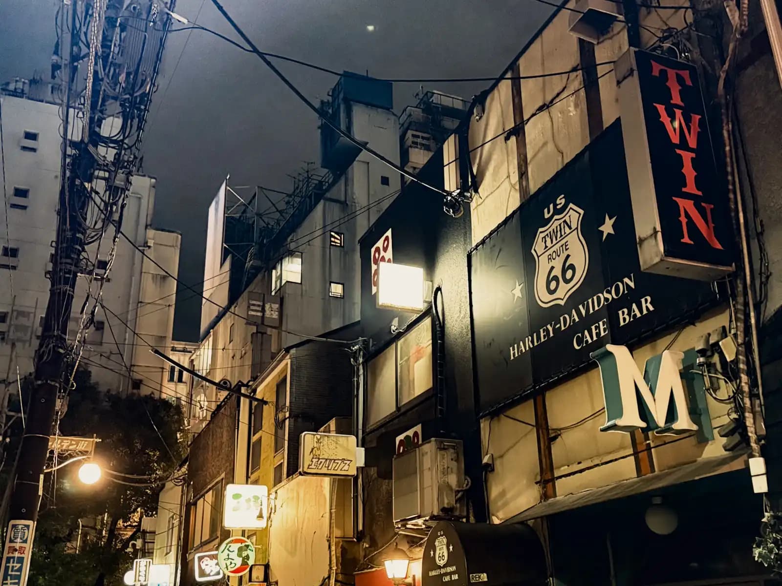 The big Twin sign for a Harley-Davidson cafe bar with a Route 66 logo, on the edge of Golden Gai under a dark sky.