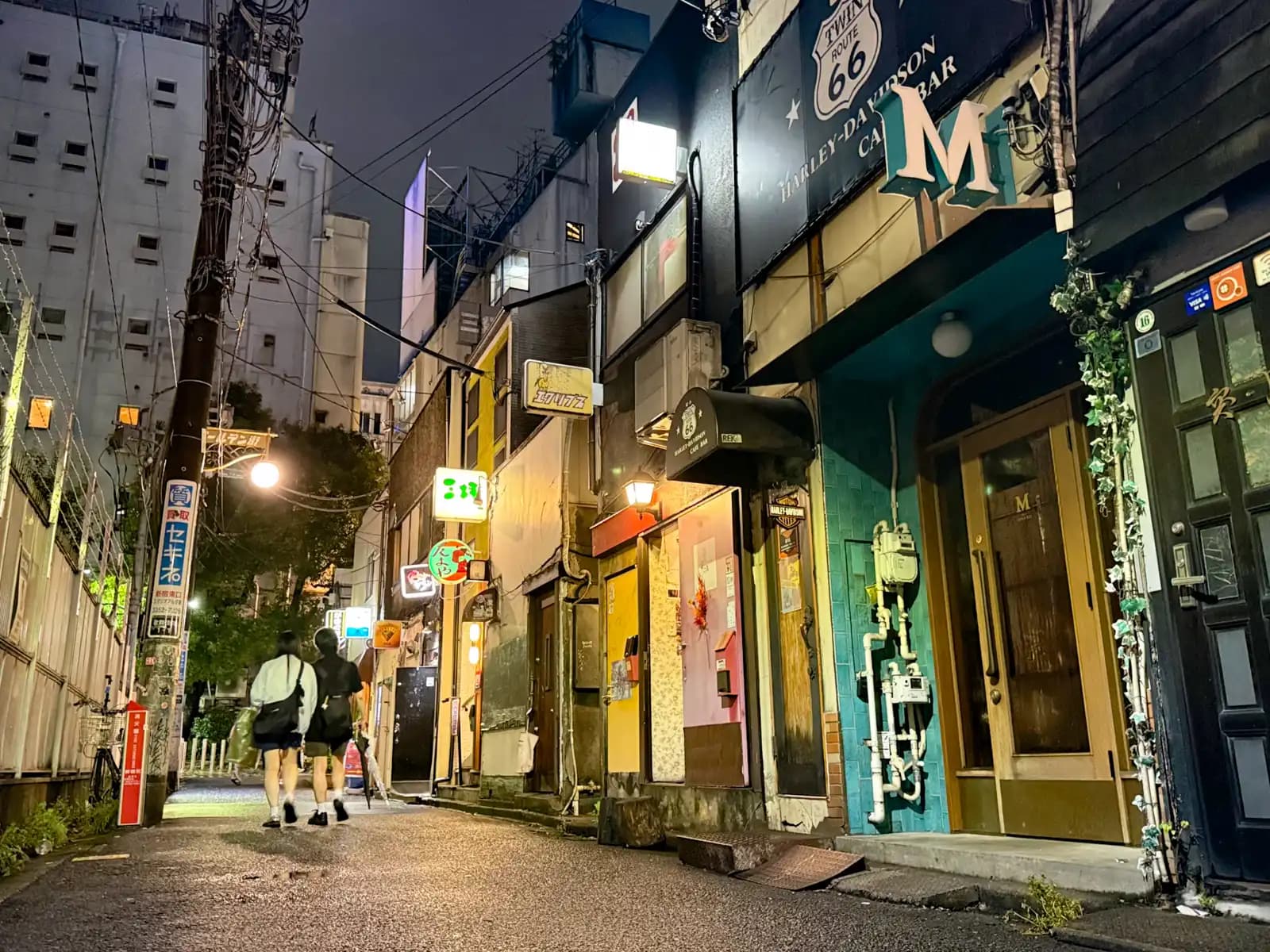 Two people with umbrellas walking along the outer street, past a teal-tiled bar front and a door covered in fake ivy.