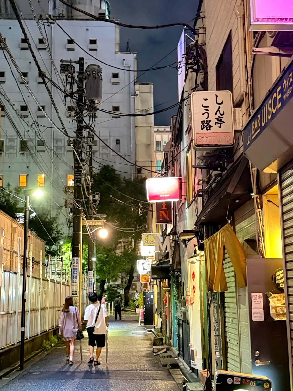 A couple walks down the outer lane past Bar Uisce and a Sapporo sign, with trees and a big building up ahead.