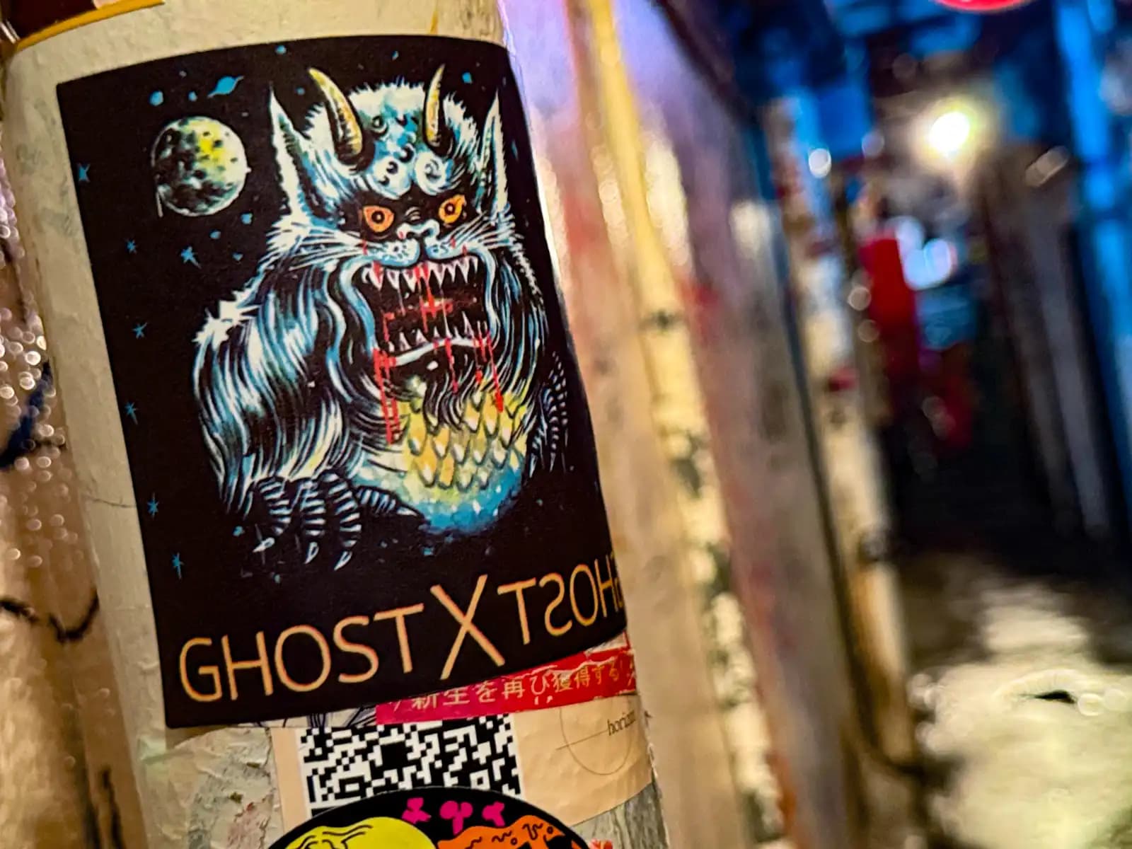 A "Ghost X Ghost" sticker of a hairy horned monster stuck on a pole, with the alley blurred behind.