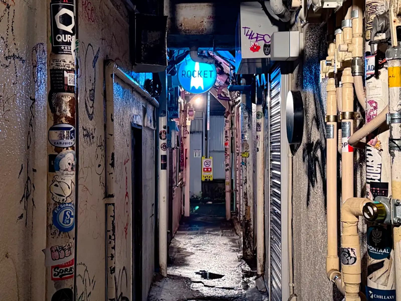 From the other end, the Rocket sign glows blue over a tight passage full of stickers, pipes and puddles.