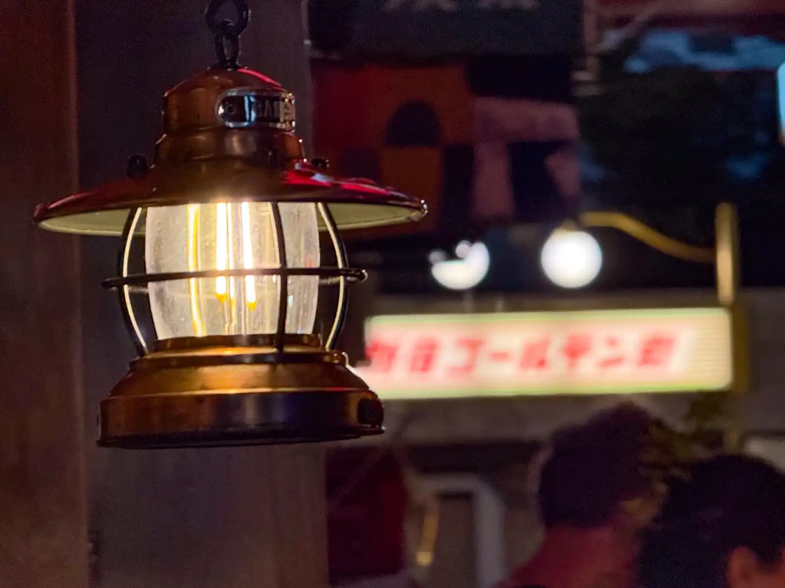A small metal lantern hanging by a bar, with the Shinjuku Golden Gai sign out of focus behind it.