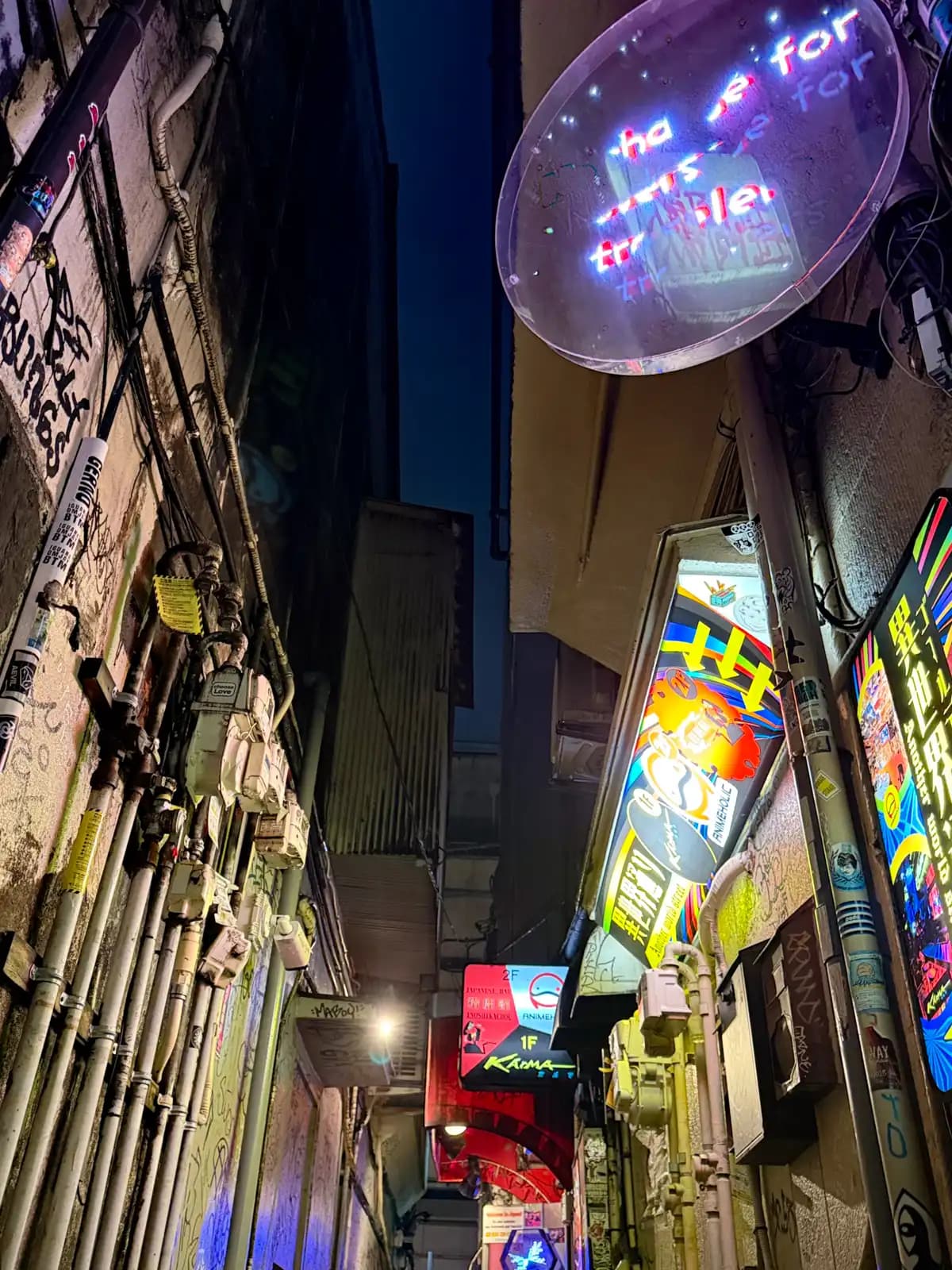 Looking up a narrow gap between buildings at a clear round neon sign, with Karma and other bright signs lower down.