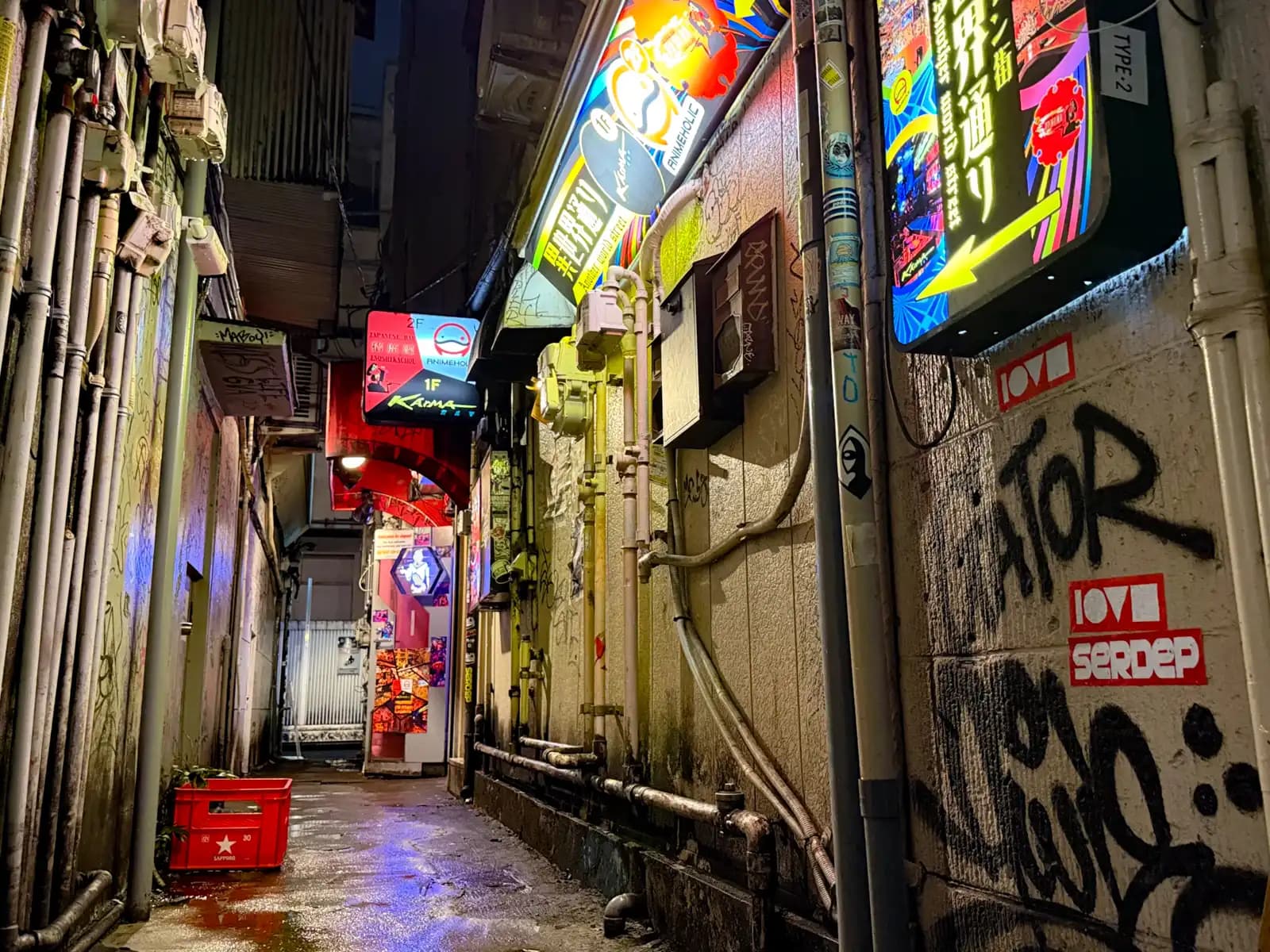 A wet back passage with the Karma sign at the end, a red Sapporo crate, pipes and graffiti on the walls.