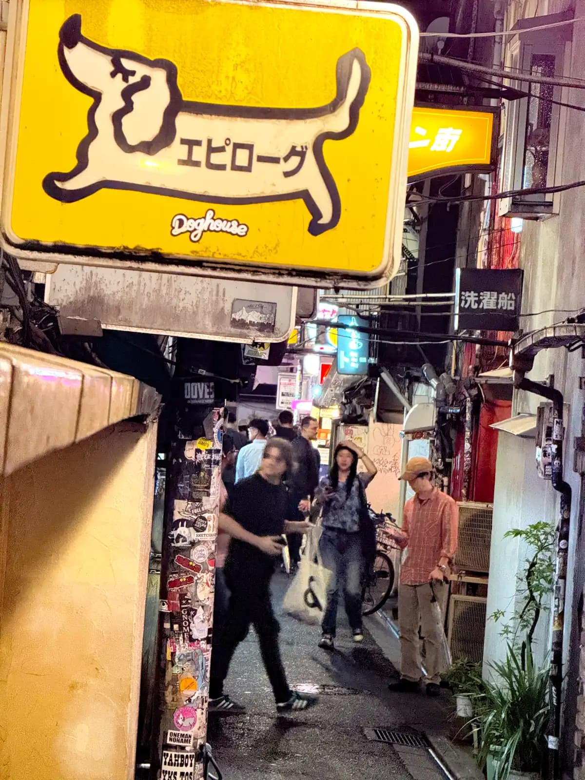 The yellow Epilogue Doghouse sign with a dachshund on it, above people walking through a busy Golden Gai lane.