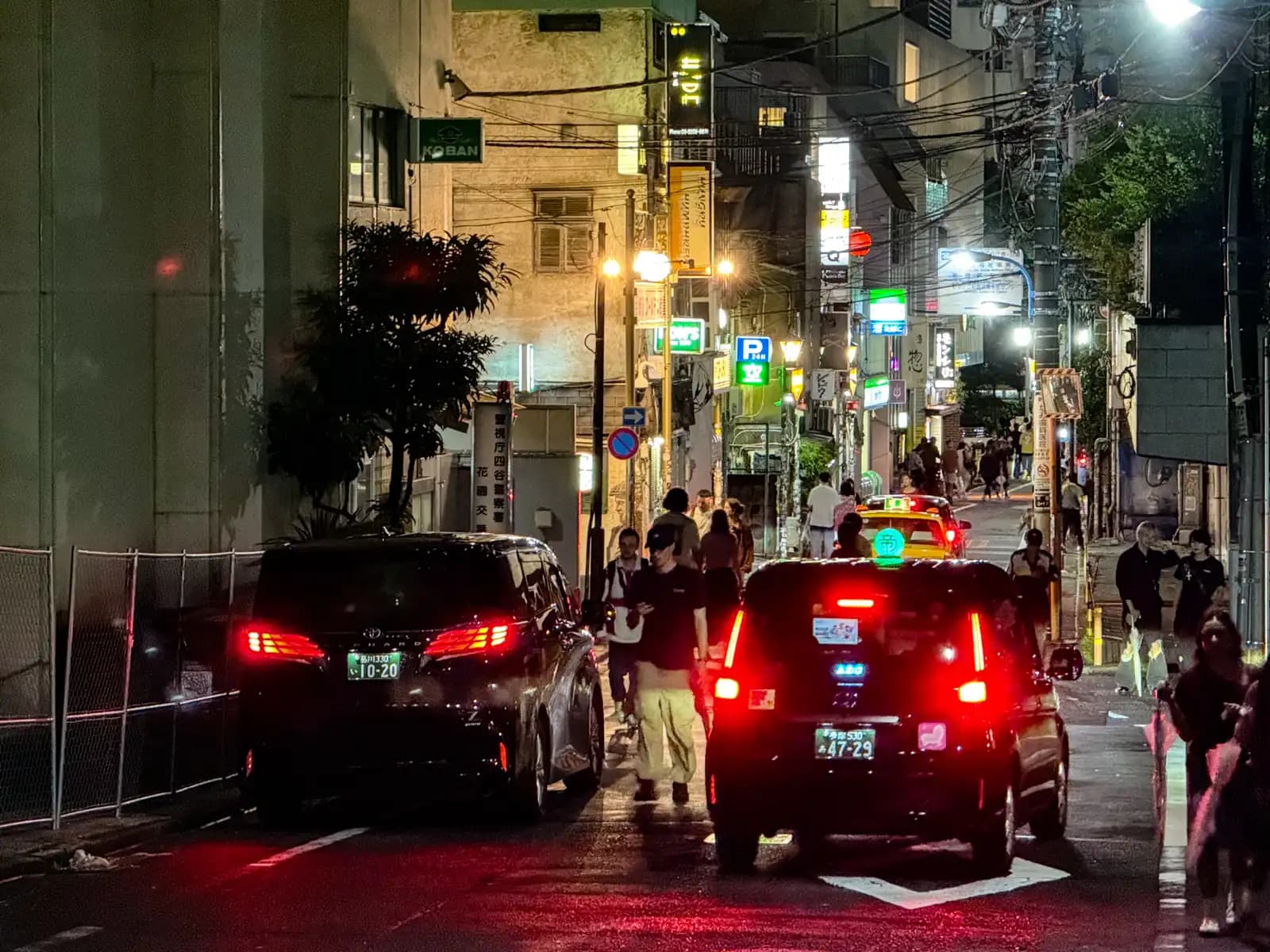 Cars and a taxi squeeze through the street just outside Golden Gai, with lots of people walking and a koban sign up on the left.
